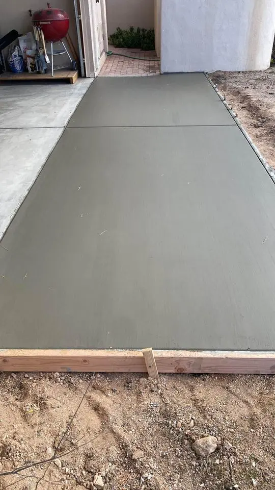 Freshly poured concrete slab drying next to a garage, framed with wooden boards on the sides, creating a seamless transition for future paver installation. A red barbecue grill is visible in the background, partially inside the garage. Dry soil surrounds this evolving outdoor space.