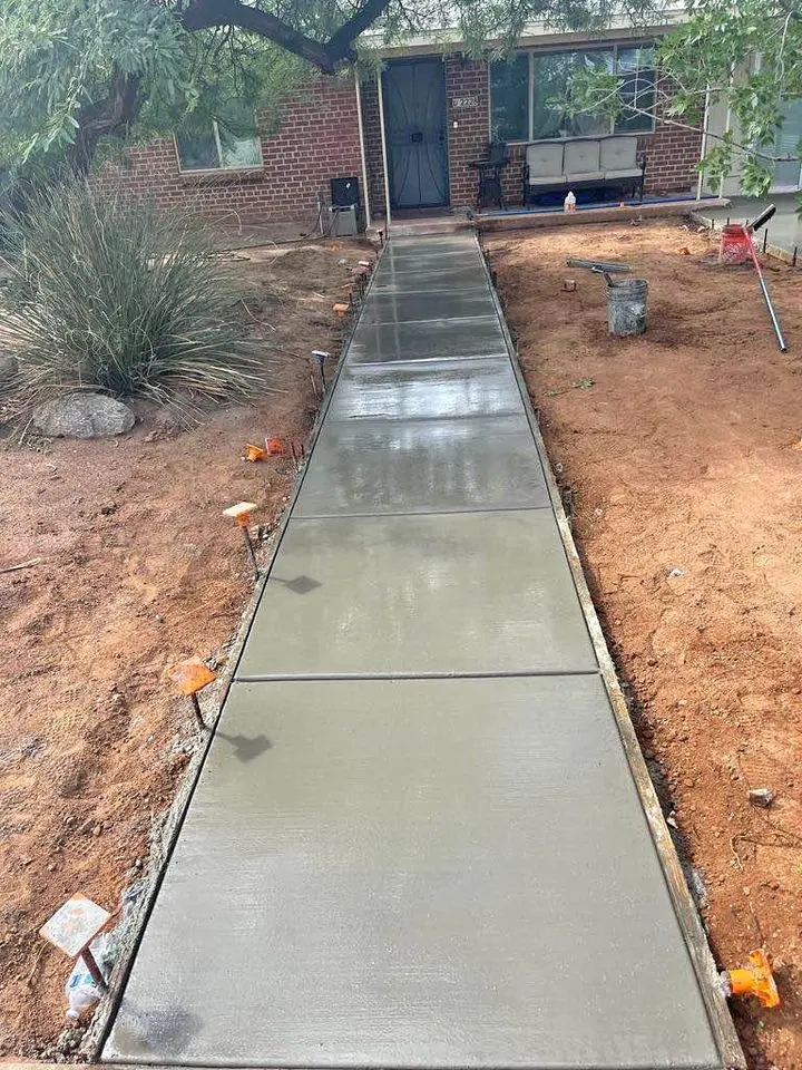 A freshly poured concrete walkway enhances the outdoor space as it leads to the front door of a brick house in Scottsdale, AZ. The path is bordered by soil and plants, with small orange markers along the edges. By the entrance, a bench invites you to pause and enjoy this tranquil setting.