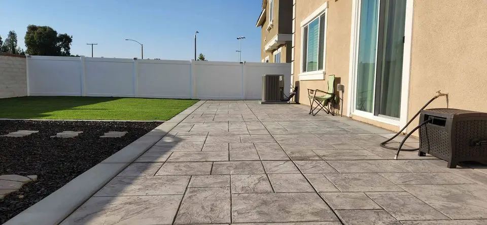 A patio with stamped concrete flooring enhances the outdoor space alongside a house with beige siding. An air conditioning unit and a chair sit by a window, enclosed by a white fence that borders the grassy area. A brown storage box is also present, perfect for those sunny Scottsdale AZ days.