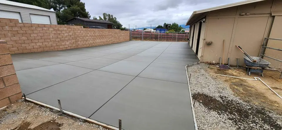 A spacious outdoor patio with a stone tile design consisting of large, irregular shapes in shades of beige, brown, and gray. The area is surrounded by dense green foliage, a small decorative round table on the left, and a clear glass sliding door on the right. Contact our licensed concrete contractors in Scottsdale AZ for a free quote today.