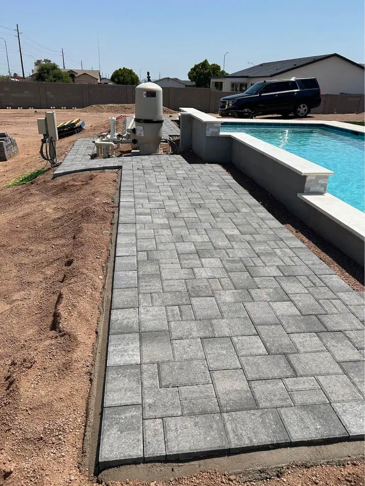 A newly installed gray stone walkway highlights the outdoor space beside a rectangular swimming pool. A black SUV is parked nearby in Scottsdale, while the surrounding area remains unlandscaped, with bare soil and some equipment visible in the background, showcasing recent paver installation.