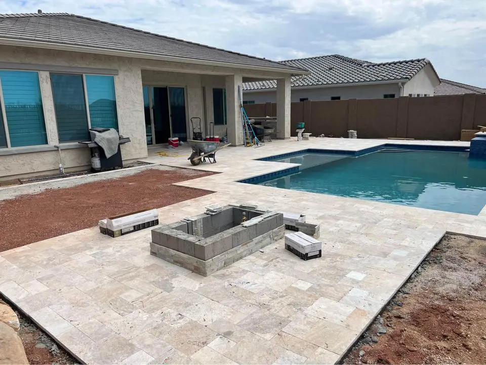 A residential backyard in Scottsdale, AZ, is under renovation, showcasing a partially completed stone patio with an in-ground pool. Paver installation materials and tools are scattered around, while the house exterior—with large windows and a covered patio area—overlooks this evolving outdoor space.