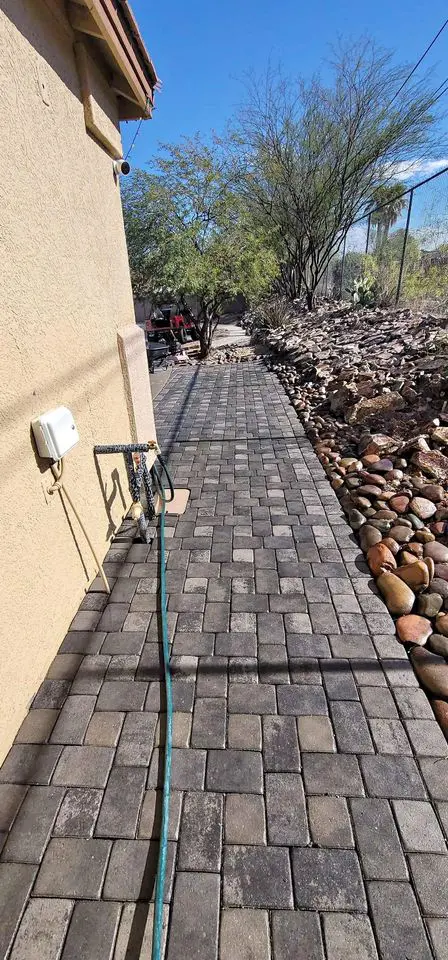 A paved walkway beside a beige building in the Phoenix area leads to a garden. On the left, a hose is stretched out, and on the right, there is a rock border with desert plants and trees under a clear blue sky.