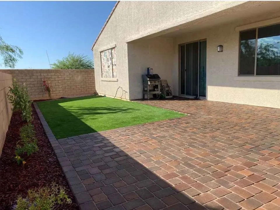 A charming outdoor space showcases a brick patio and a small artificial grass lawn, perfect for relaxation. The barbecue grill sits snugly against the house wall. Enclosed by a brick fence, this Scottsdale, AZ backyard features lush plants lining the garden bed for added serenity.