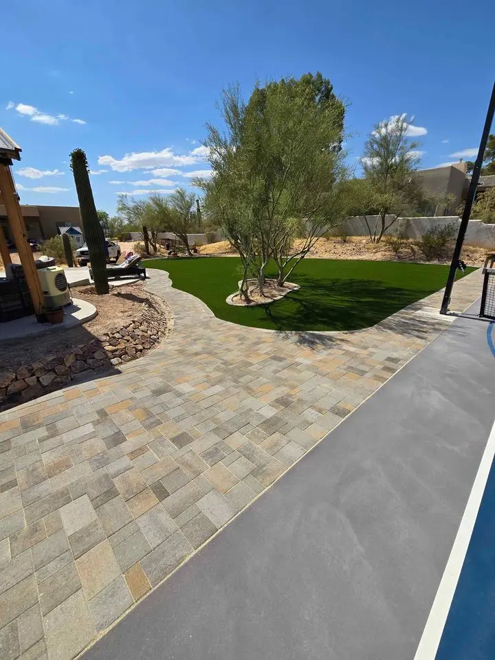 Backyard with a stone path, green artificial grass, and desert landscaping adorned with cacti. This inviting outdoor space features a swimming pool edge in the foreground and trees lining the background. The sky remains clear and blue, reminiscent of Scottsdale, AZ's serene ambiance.