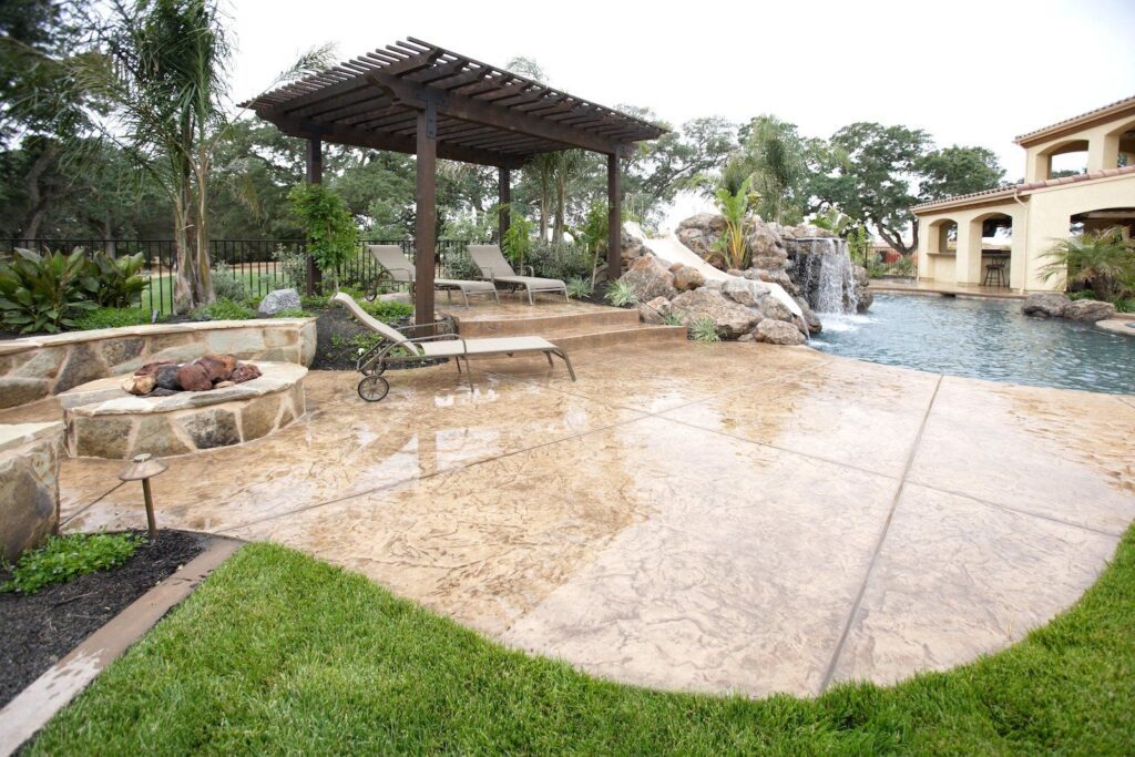 A luxurious backyard in Scottsdale AZ features a swimming pool with a rock waterfall. A wooden pergola stands over lounge chairs on a concrete pool deck by Scottscrete Contracting. A fire pit and lush greenery complete the scene, providing a serene and inviting outdoor space.