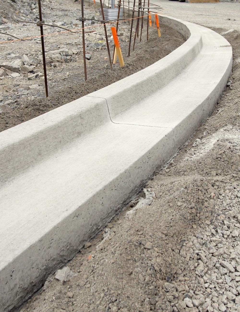 Freshly constructed concrete curb in a gravelly construction area, partially fenced with rebar and caution tape in the background. The curb gently curves around, following the road's edge. Built by licensed concrete contractors in Scottsdale AZ, this project ensures quality and durability. Contact us for a free quote!