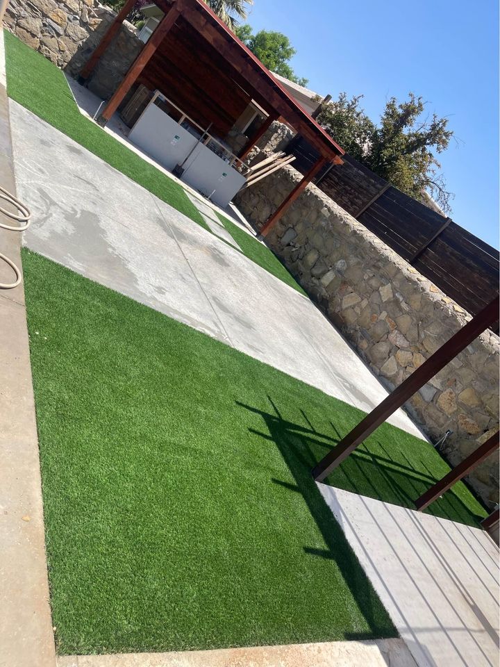 A backyard area with artificial green grass and concrete walkways. A wooden pergola structure in the back corner partly covers an outdoor kitchen with a grill and a sink on a concrete slab. The yard, enclosed by a stone wall, is perfect for enjoying the clear and sunny Scottsdale, AZ sky.