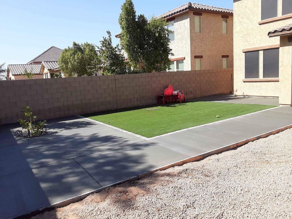 A backyard with a concrete patio surrounds a rectangular patch of artificial grass. Red lawn chairs are placed at the far end of the grass area. The space, expertly crafted by a Scottsdale concrete contractor, is enclosed by a brick wall, with neighboring houses visible over it and a small tree in one corner.