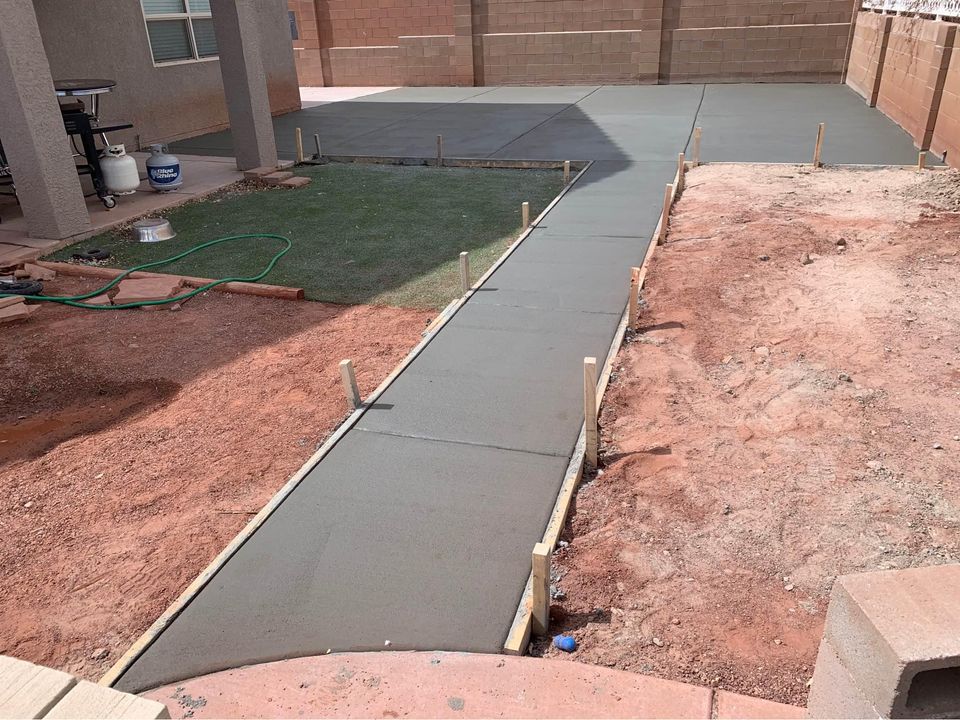 A backyard under construction in Scottsdale AZ features newly laid concrete slabs. The path runs from the foreground to a patio area in the background, bordered by red soil. Patches of green grass, a garden hose, and a BBQ grill near the house complete the scene.