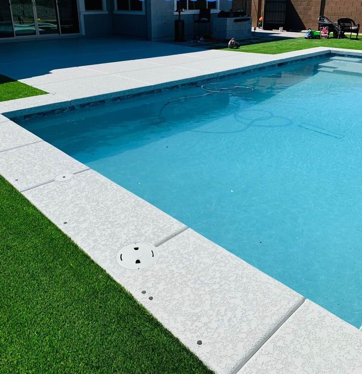 A clear blue swimming pool with a concrete deck, expertly crafted by Scottscrete Contracting, is surrounded by lush artificial grass. The pool area in this Scottsdale AZ backyard features outdoor furniture, including a table with chairs, all brightly lit by sunlight.