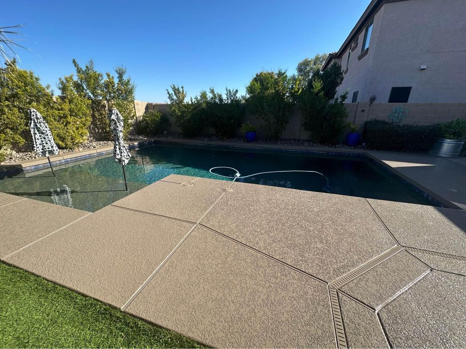 A backyard swimming pool with clear water is surrounded by a concrete deck designed by Scottscrete Contracting. Two umbrellas with black and white patterns stand on the pool's shallow end. Bushes and trees line the fence in the background, with a portion of a house visible on the right.