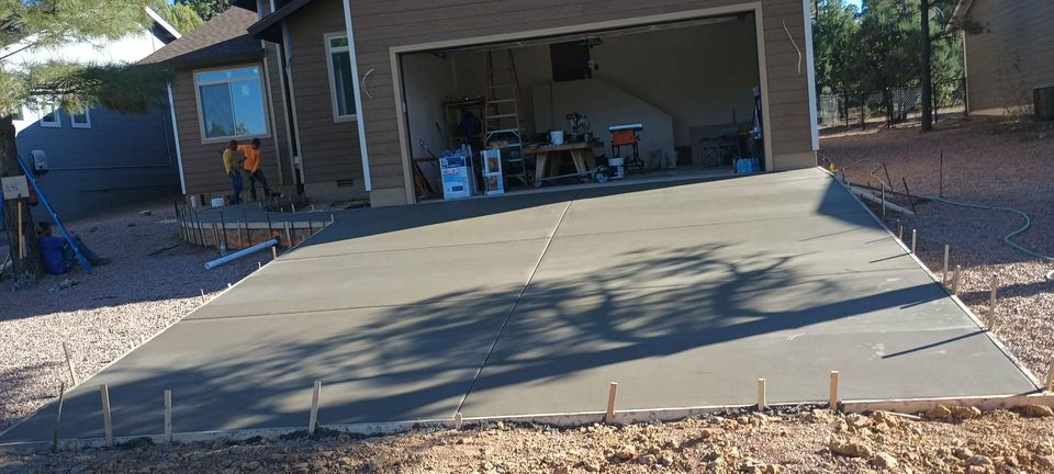 A newly poured concrete slab driveway in front of a garage is in the process of setting. The garage is open and contains various tools and equipment. A person is working near the side of the house. The area around the driveway, which was laid by a foundation contractor from Scottsdale AZ, is unpaved and rocky.