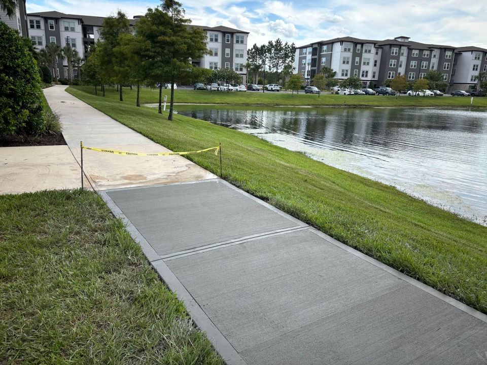 A freshly laid concrete sidewalk section, crafted by Scottsdale concrete contractors and roped off with yellow caution tape, runs alongside a grassy area near a pond. Modern apartment buildings are visible across the green space. The sky is partly cloudy.