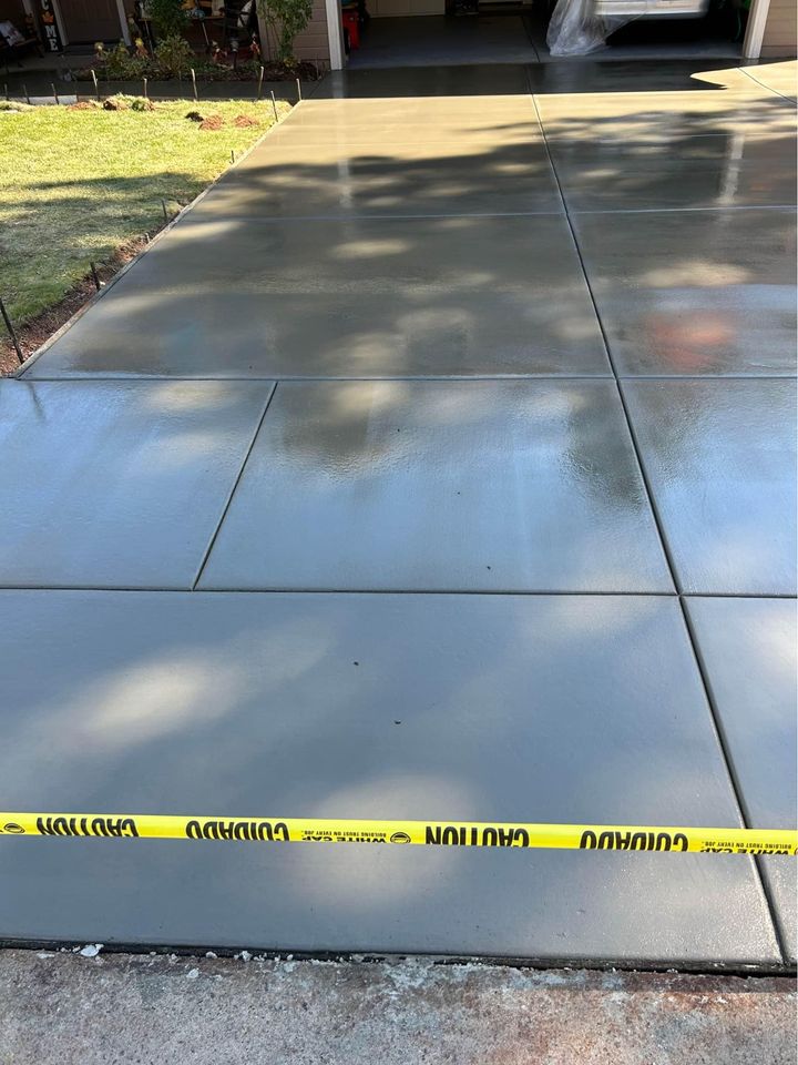 A freshly poured concrete driveway installation in Scottsdale, AZ, sectioned into squares, reflects light from its smooth, wet surface. A yellow caution tape runs across the entrance to prevent access. A garage and some landscaping are visible in the background.