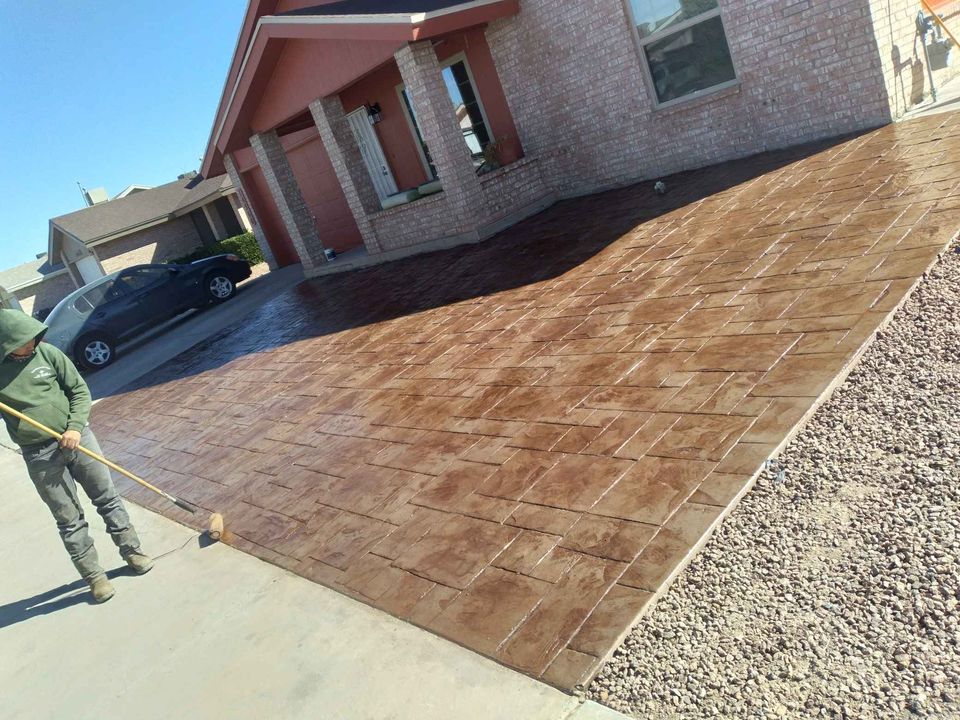 A worker in a green hoodie and jeans sweeps a newly installed brown textured driveway in front of a brick house with a paved porch. A black car is parked nearby and there's a gravel section on the side, showcasing the meticulous work of Scottsdale concrete contractors.