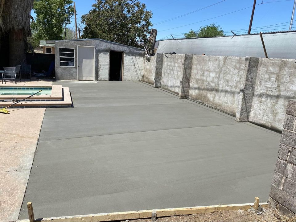 Freshly poured concrete slab in a backyard next to a swimming pool in Scottsdale, AZ. The smooth, grey surface extends to the stone wall surrounding the area. Completed by licensed concrete contractors, there's a small building with a door and window at the far end and some chairs visible near the pool.