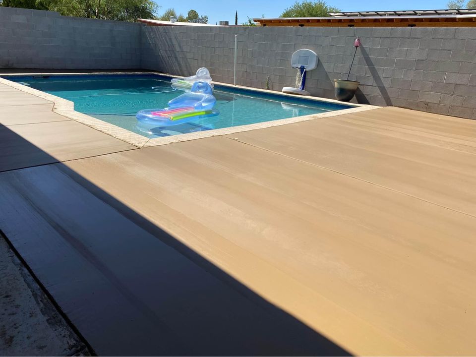 A backyard swimming pool with a cement deck, courtesy of Scottscrete Contracting in Scottsdale AZ, and a gray block wall surrounding it. A clear inflatable pool float with a rainbow design is floating in the water. There is also a basketball hoop attached to the wall at the pool's edge.