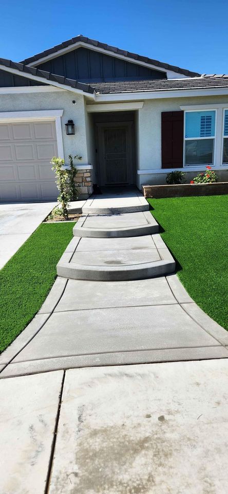 A modern single-story house in Scottsdale, AZ, with a gray roof and white exterior. The front entrance features a series of curved concrete steps installed by licensed concrete contractors, flanked by well-manicured, lush green artificial grass. The garage door is on the left, and there are decorative plants near the door.