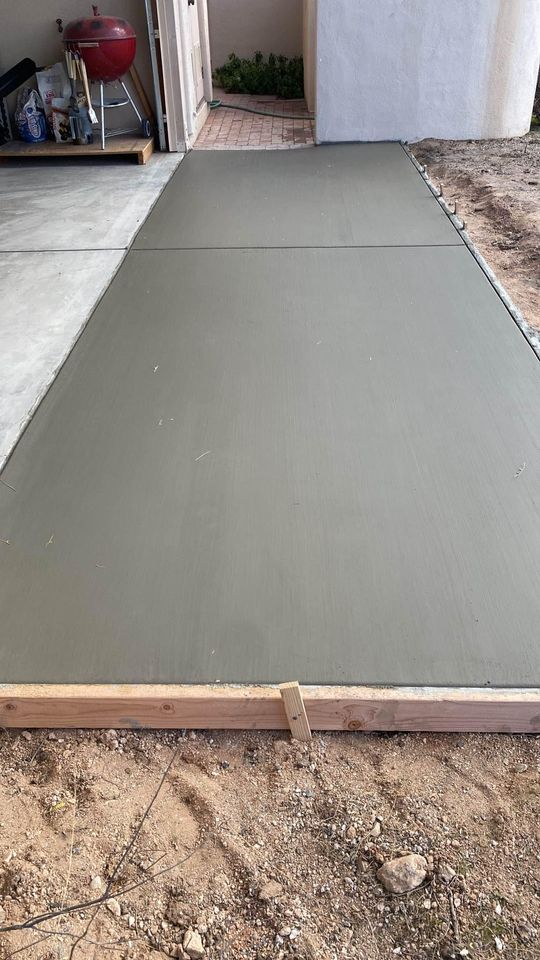 A freshly poured concrete slab, crafted by licensed concrete contractors in Scottsdale AZ, extends from a garage to an outdoor area. The wet concrete is bordered with wooden frames, and a small clip is fastened to the form at the bottom. In the garage, a red grill is visible to the left.