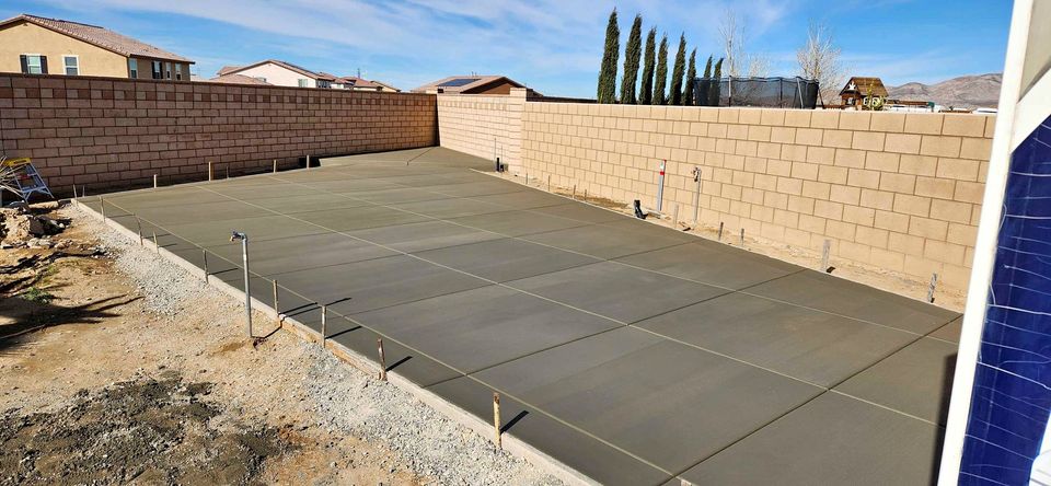 A freshly poured concrete slab in a Scottsdale backyard, enclosed by a high brick wall. The ground around the concrete is a mix of dirt and gravel. Residential houses and trees are visible in the background under a clear, blue AZ sky; ideal work by local concrete contractors specializing in driveways.