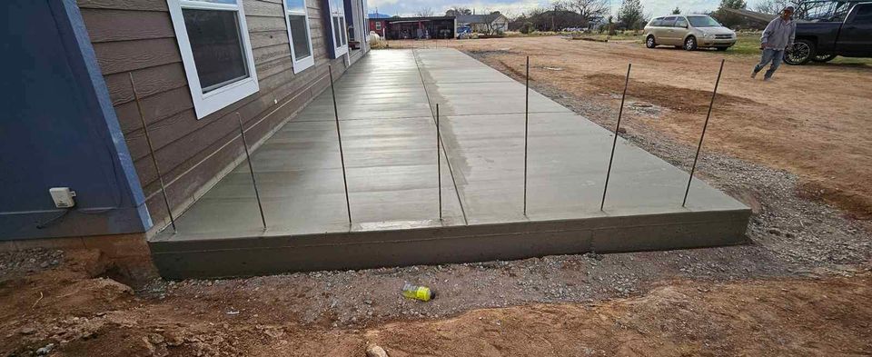 A freshly poured concrete slab extends from the side of a building with brown siding and white-framed windows. Metal rods are embedded in the slab by a Scottsdale concrete contractor. The surrounding area is a dirt construction site, and a person walks in the background. Ask about our free concrete service quote!