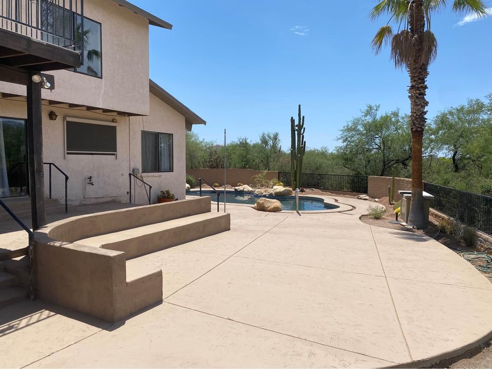 A backyard in Scottsdale, AZ features a curvy in-ground pool surrounded by desert landscaping, including rocks and a large Saguaro cactus. There is a beige stucco house with railing leading to steps and lush trees in the distance under a clear blue sky, thanks to Scottscrete Contracting's expertise.