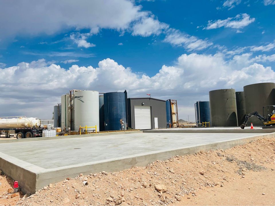 An outdoor industrial area under a blue sky with clouds features large cylindrical storage tanks, a small building with a white door, and a smooth concrete slab in the foreground laid by an Arizona concrete contractor. A truck is parked alongside the tanks, and a construction vehicle is also visible in the scene.