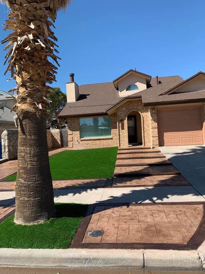 A two-story house with a brick exterior and brown roof stands on a sunny day in Scottsdale, AZ. The front yard features a neat green lawn with a large palm tree and a paved walkway leading to the garage and front door. Considering concrete driveway installations? Contact us for a free quote!