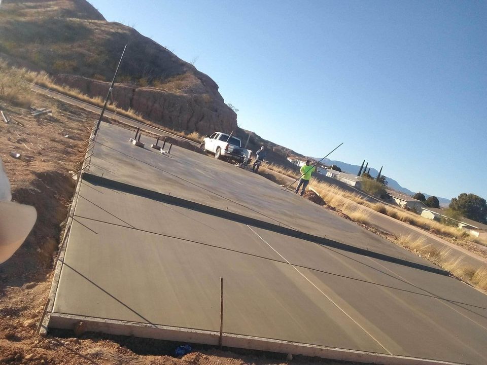 A construction worker in a green shirt and hard hat smooths out a large concrete slab foundation at a construction site in a rural area. A white truck is parked nearby, with rocky terrain and clear blue skies in the background—courtesy of your reliable commercial concrete contractor.