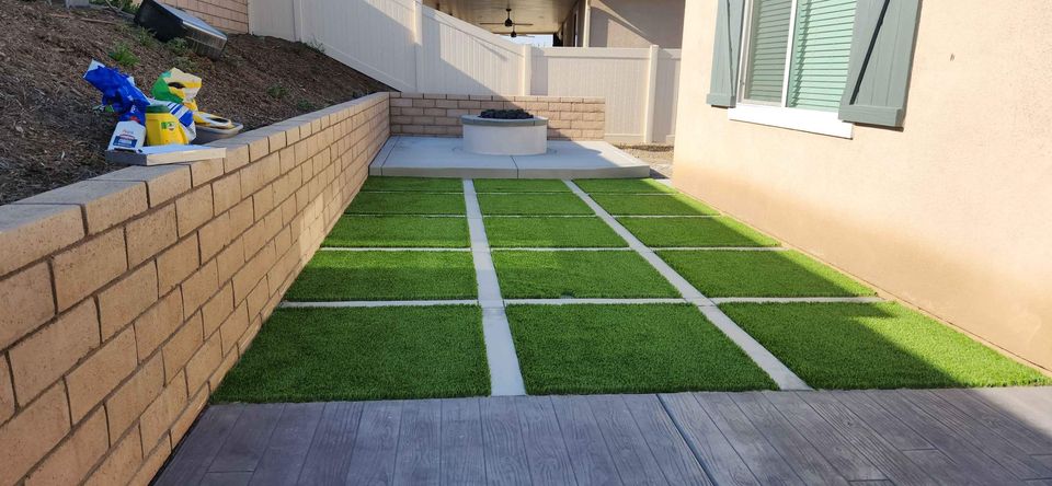 A backyard area in Scottsdale AZ with a grid pattern of artificial grass sections separated by concrete tiles. On one side, there is a brick retaining wall and on the other, the exterior wall of a house with green shutters. Gardening supplies are on the retaining wall. Licensed concrete contractors offer a free quote for enhancements.