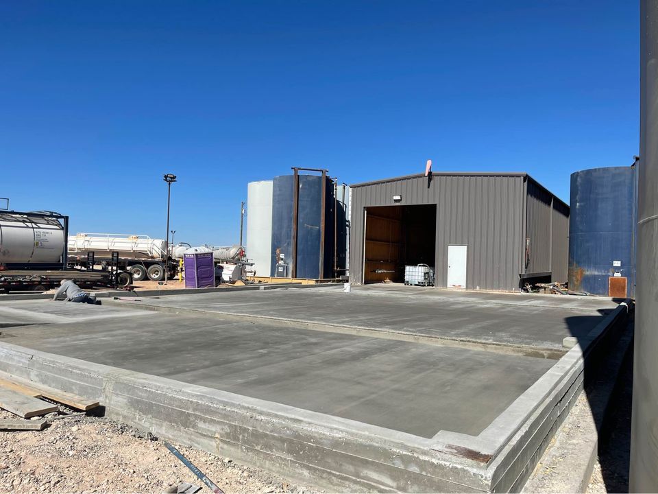 A sunny outdoor scene depicts a construction site with a large, newly poured concrete foundation in the foreground. In the background, there is a metal shed with its door open, several storage tanks, and parked trucks. The Scottsdale concrete contractor oversees the project on dry, barren land.