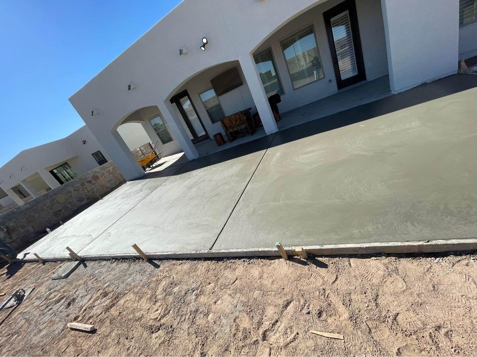 A newly poured concrete patio extends from a white stucco building with arched openings and several doors and windows. The foreground shows an unpaved, sandy area. The patio, expertly crafted by a Scottsdale concrete contractor, is bordered by a stone wall on the left side. The sky is clear and sunny.